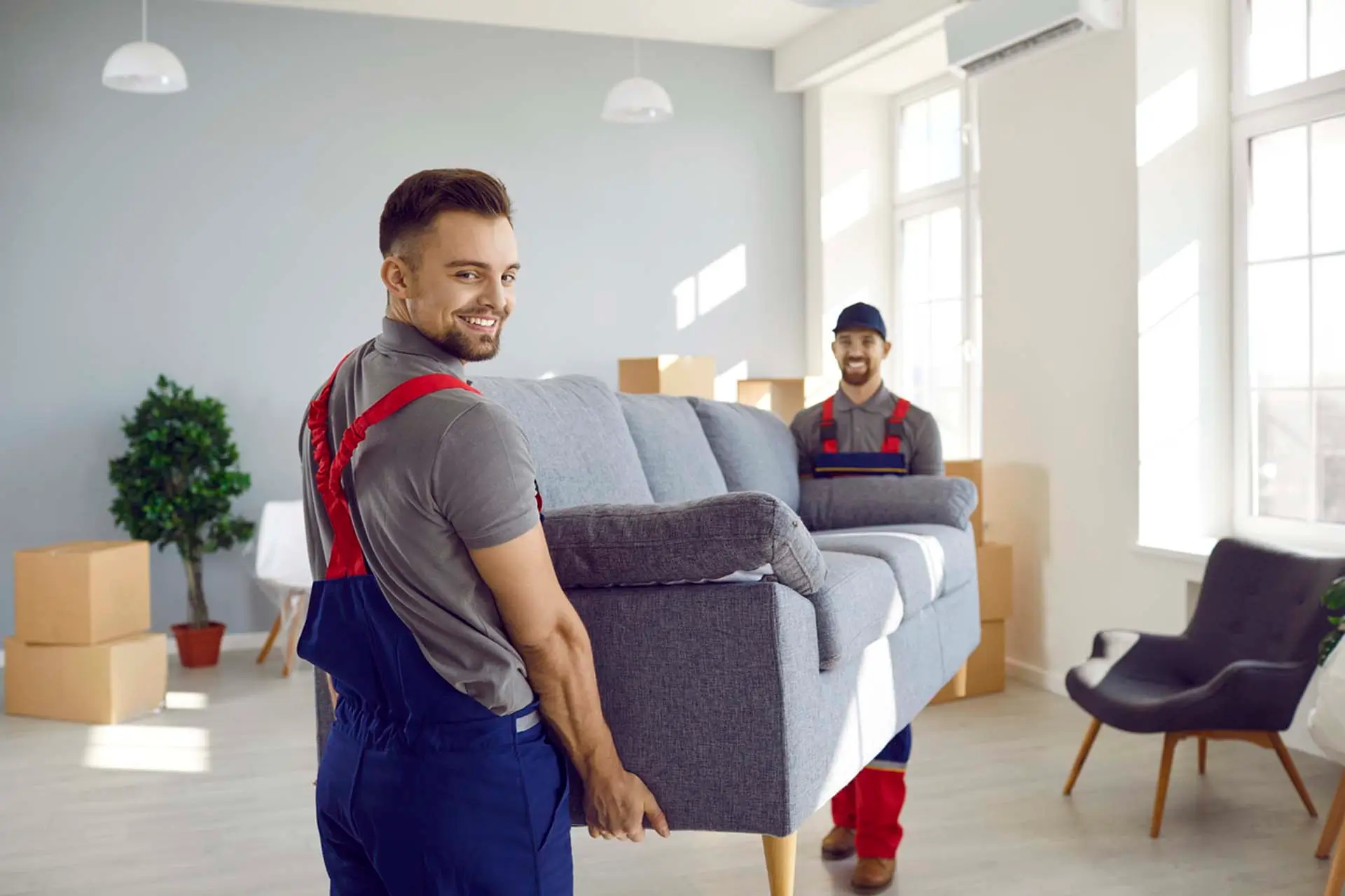 Zwei Smile-Umzugshelfer in Uniform tragen ein graues Sofa in ein helles Zürcher Zimmer mit Kisten und Pflanzen.